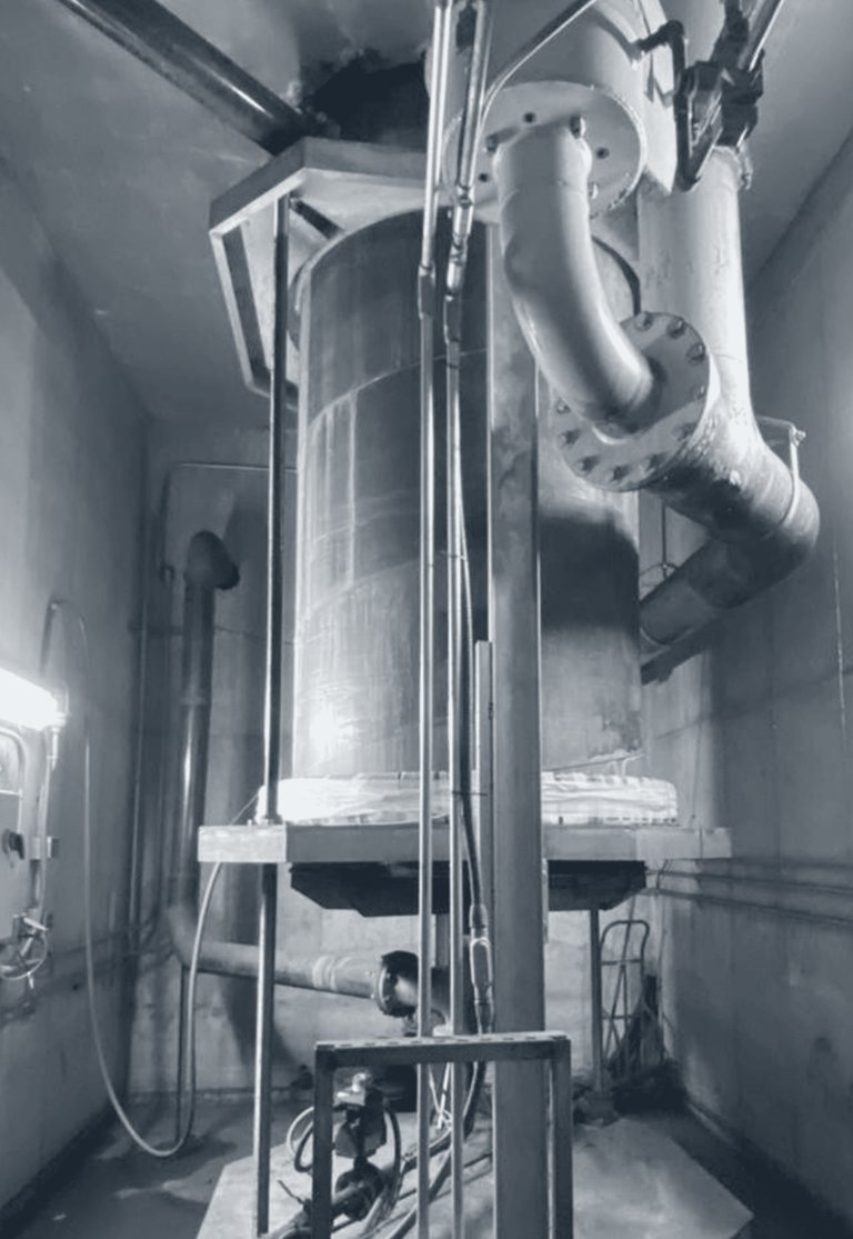Photograph of a crucible in a furnace pit with a carriage plate part way down, vertical guide rods and a drive at the base.