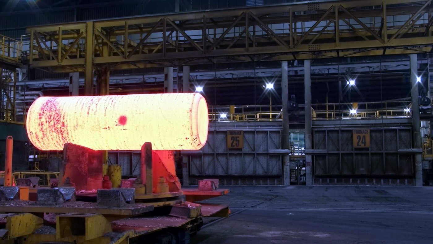 Wide photograph of a glowing orange billet on press tooling in a large forge building with structural steel and furnace doors behind it.
