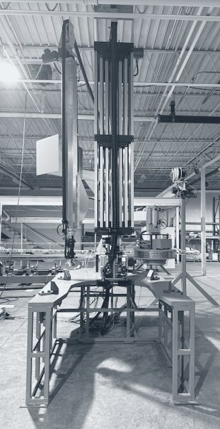 Photograph of a tall scanner rig in a workshop: a vertical telescoping mast above a steel base frame, with a crucible section on a stand beside it.