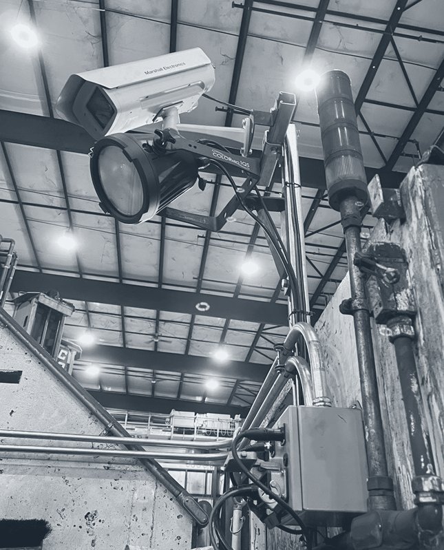 Photograph of a camera in a shielded housing on a mast at a furnace, with a light, conduit and a junction box below.